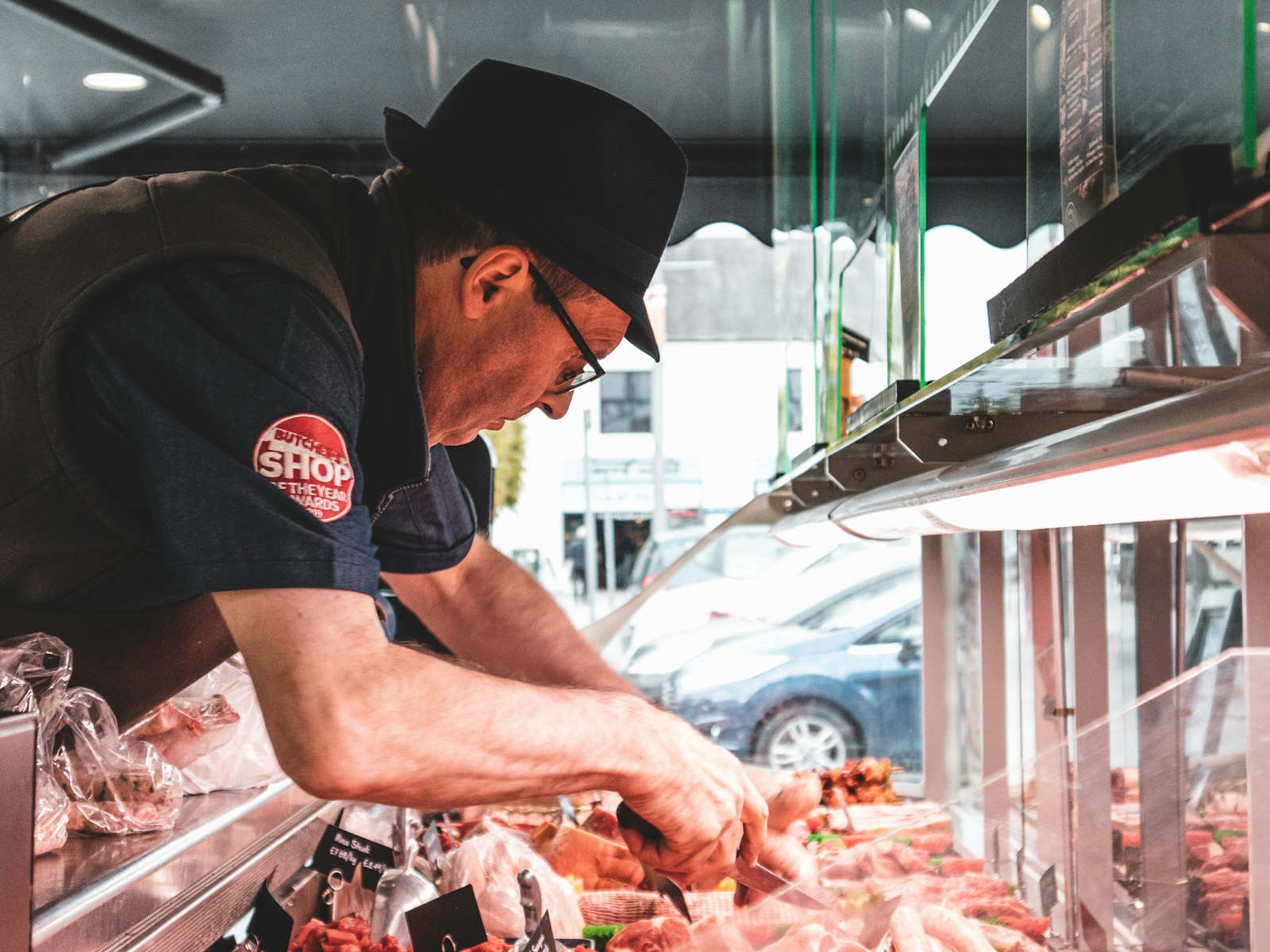 Artisan butcher in a flat cap carefully arranging cuts in his shopfront display case